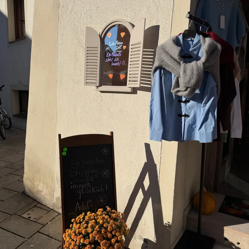 Ein kleines Geschäft mit einem Kreidetafelschild und Blumen in einem Topf. Das Schild sagt 'Ein Besuchen Shopping macht immer glücklich!'. Es hängen Kleidungsstücke an der Wand und ein Fenster mit einem Herzdesign.