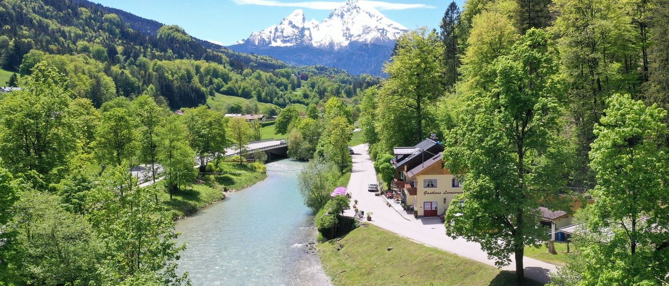 Luftaufnahme eines Flusses mit einem gelben Gebäude rechts. Berge mit Schnee in der Ferne.
