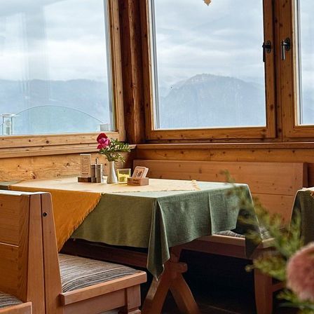 Ein rustikaler Essbereich mit Holzwänden und Fenstern. Ein Tisch hat eine grüne Tischdecke und eine Vase mit einer Blume. Die Aussicht draußen zeigt Berge und einen bewölkten Himmel.