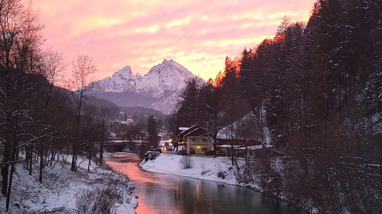 Eine friedliche Winterlandschaft mit einem Fluss unter einem farbenfrohen Sonnenuntergang, schneebedeckten Bergen im Hintergrund und einem Haus am Flussufer.