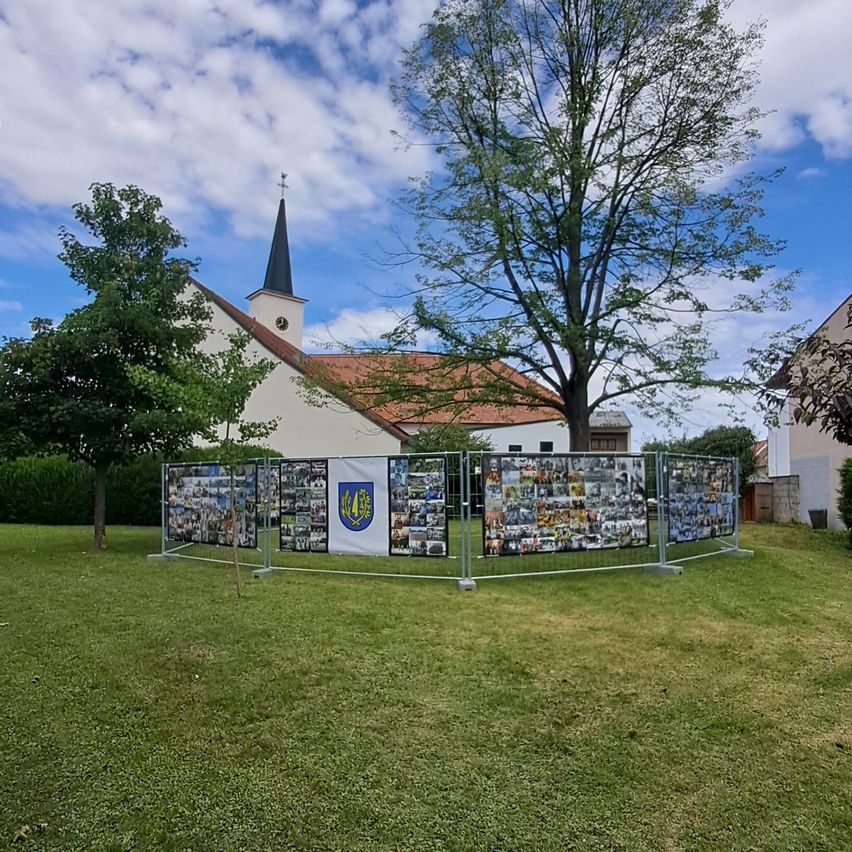 Eine Kirche mit einem Kirchturm und einem roten Dach ist von einem Metallzaun mit Collagen umgeben. Grüner Rasen und Bäume sind im Vordergrund.