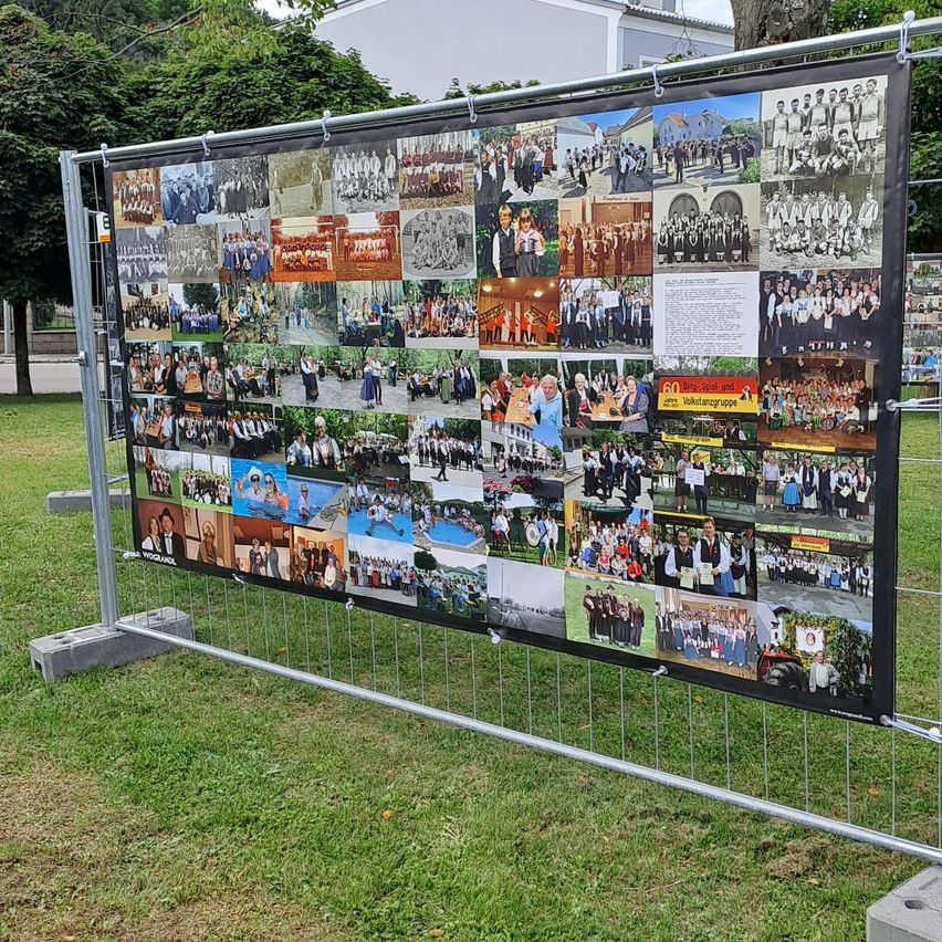 Ein großes Banner mit zahlreichen Fotos wird auf einem Metallzaun in einem Park ausgestellt. Die Fotos zeigen Gruppen von Menschen in verschiedenen Umgebungen.