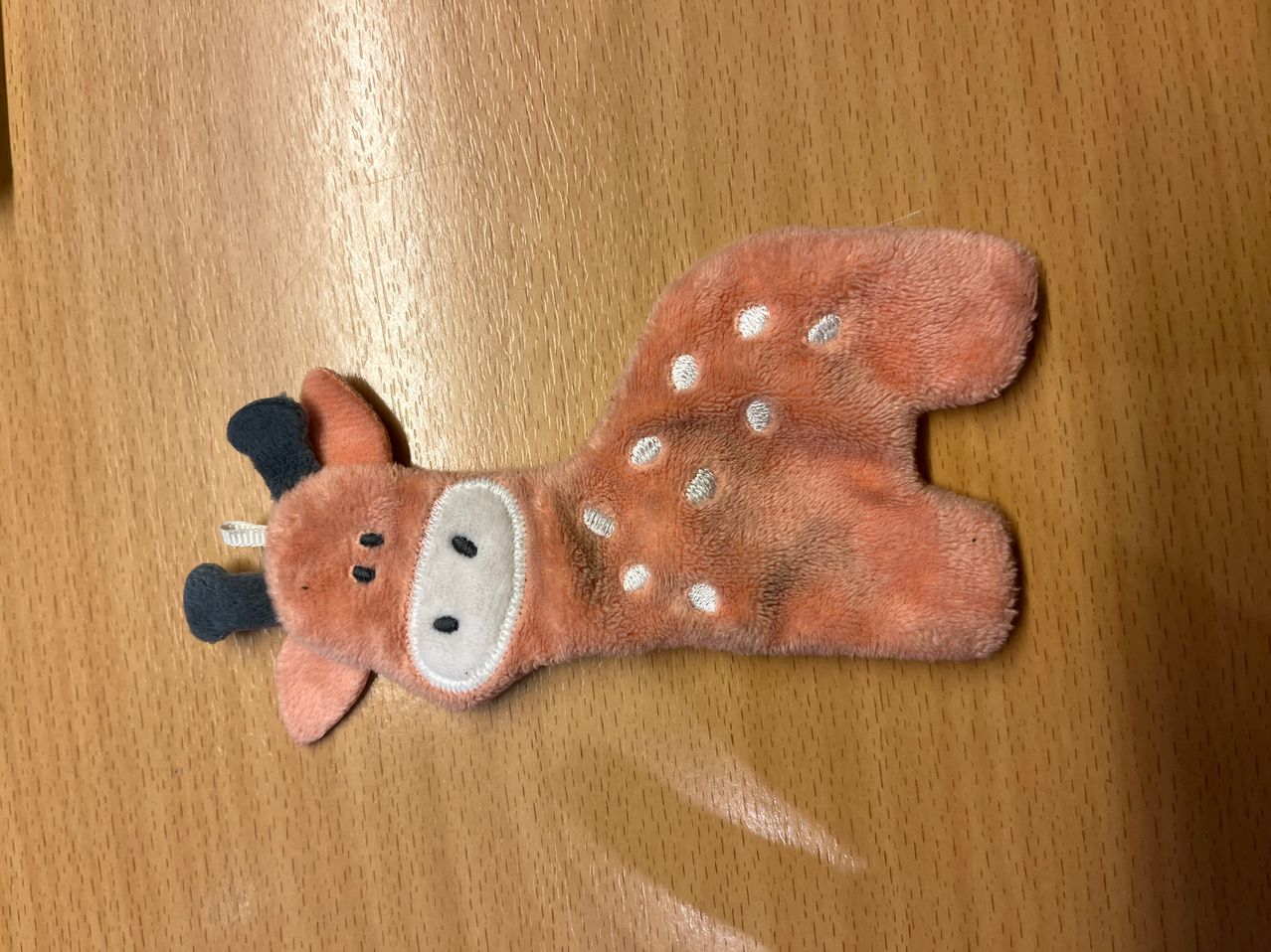 Ein brauner Plüsch-Giraffe mit weißen Punkten liegt auf einer Holzoberfläche. Das Spielzeug hat schwarze Ohren und einen schwarzen Schwanz.
