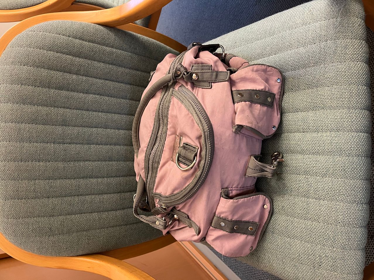 A pink backpack is placed on a wooden chair. The chair has gray cushions. The backpack has several zippers and pockets.