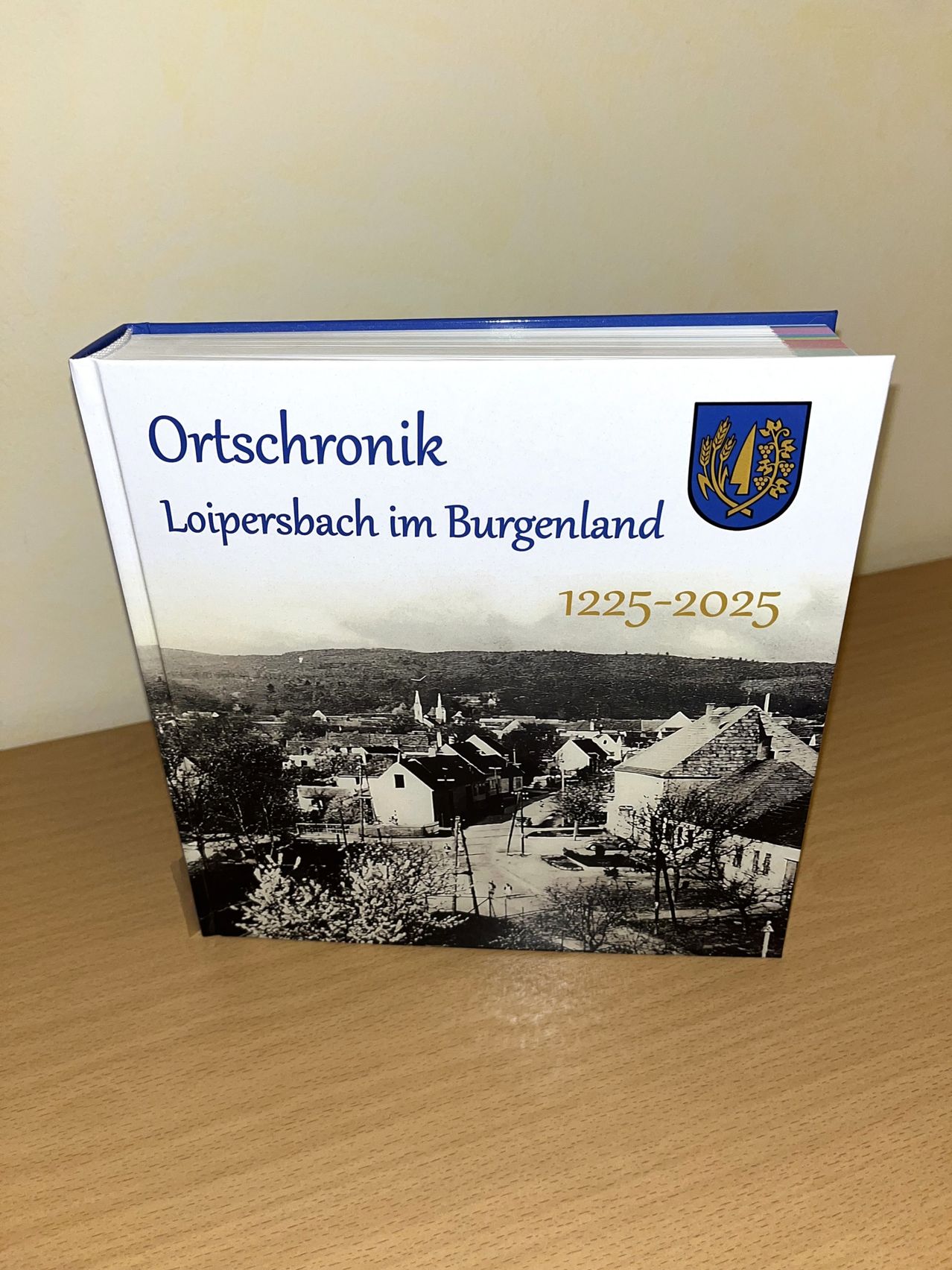 Ein Buch mit dem Titel 'Ortschronik Loipersdorf im Burgenland' wird auf einem Holztisch ausgestellt. Der Buchdeckel zeigt ein Schwarz-Weiß-Bild eines Dorfes, und der Titel enthält die Jahre 1225-2025.