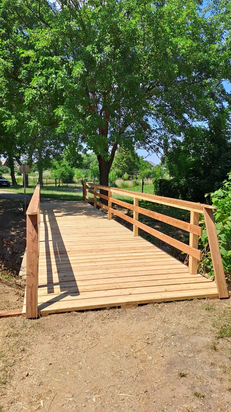 Eine hölzerne Brücke mit Geländern, umgeben von Bäumen und Gras. Ein Auto ist in der Ferne geparkt.