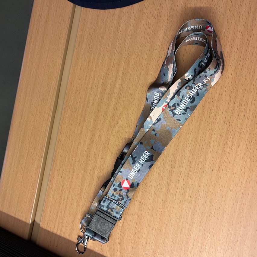 Ein Tarnlanyard mit 'Bundesheer' in Weiß auf einer Holzoberfläche. Der Lanyard hat ein rotes Dreieck mit einem Pfeil.