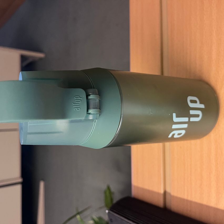 Ein grüner Thermosbehälter mit der Aufschrift 'air up' ist an einer Holzwand befestigt. Der Thermosbehälter hat einen durchsichtigen Kunststoffgriff und ein silbernes Metallelement.