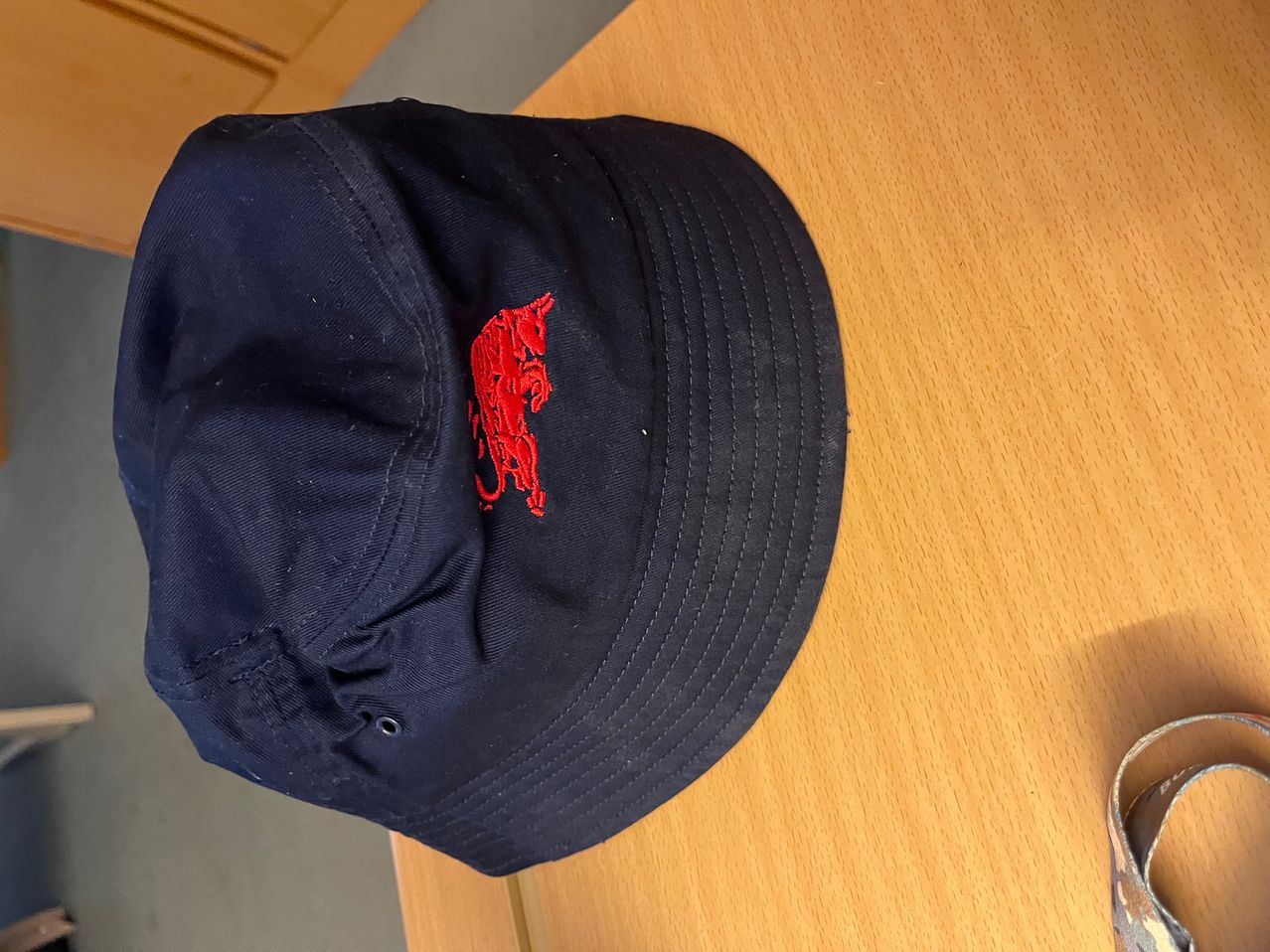 Ein schwarzer Baseball-Cap mit rotem Drachenlogo ist auf einer Holzoberfläche zu sehen. Der Drache ist schräg aufgestickt.