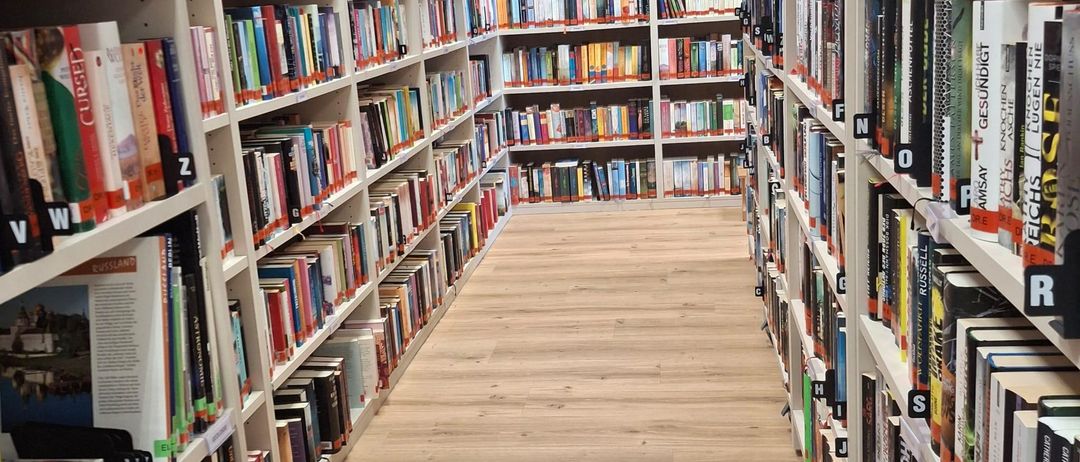 Ein Bibliotheksgang gefüllt mit Büchern auf weißen Regalen, beschriftet mit Buchstaben und Zahlen, und einem Holzboden.