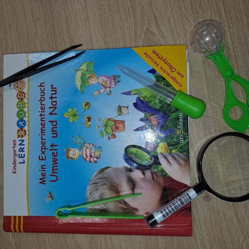 Ein Kinderbuch mit dem Titel 'Mein Experimentierbuch Umwelt und Natur' liegt auf einer Holzoberfläche. Das Buch wird von einer Lupe, Pinzetten und einer Lupe mit grünem Griff begleitet.