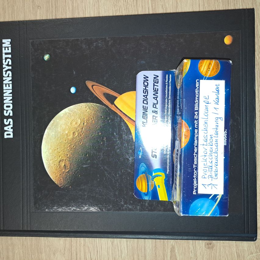 Ein schwarzes Buch mit dem Titel 'Das Sonnensystem' mit einem Bild des Sonnensystems auf dem Cover, das Planeten und Monde zeigt, zusammen mit einem Projektorfolienpaket.