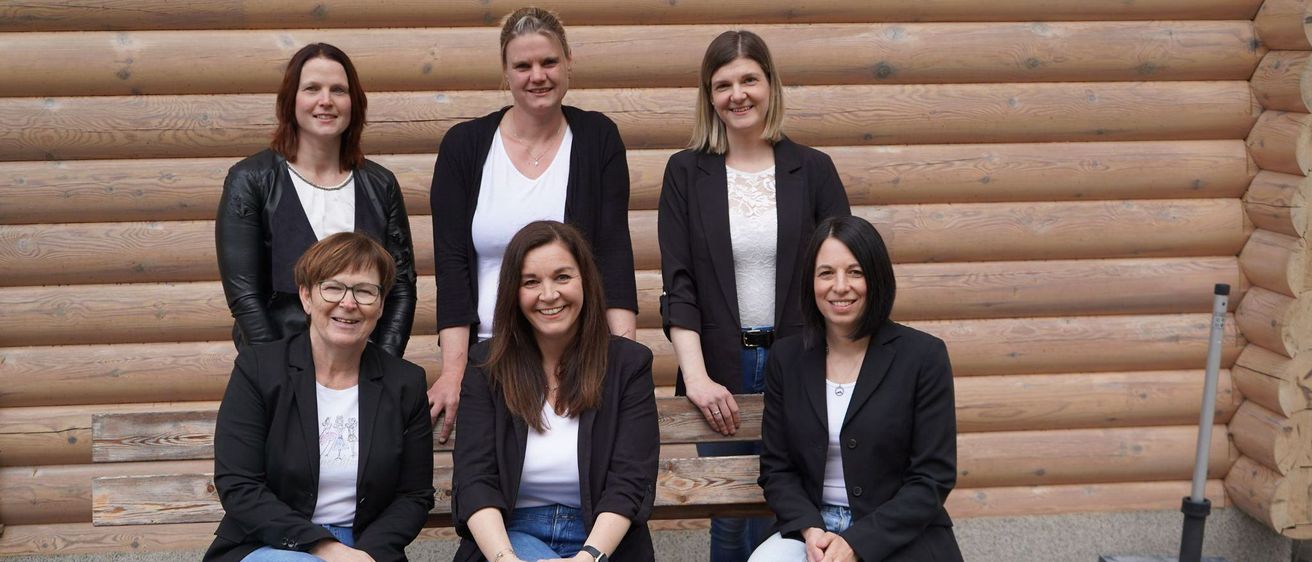 Sechs Frauen, gekleidet in schwarze Blazer und weiße Hemden, sitzen auf einer Holzbank vor einer Holzwand. Sie alle lächeln und scheinen für ein Foto zu posieren.
