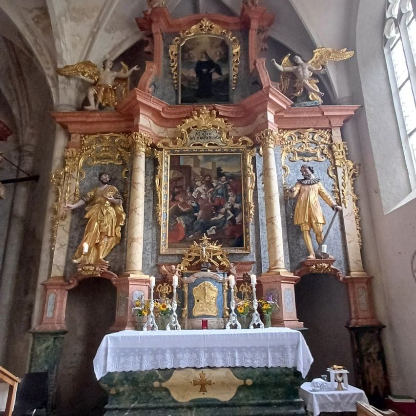 Ein kunstvoll verzierter Altar in einer Kirche mit goldenen Statuen, Gemälden und Kerzen. Der Altar ist zentriert mit einem großen Gemälde darüber und zwei Statuen auf jeder Seite.