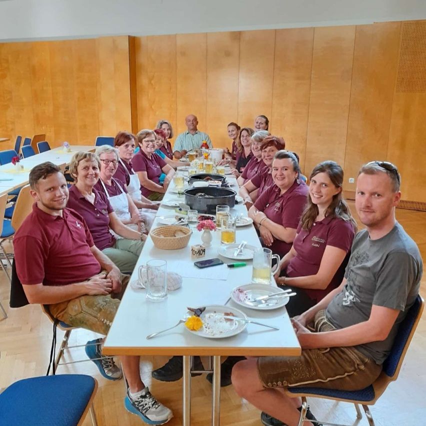 Eine Gruppe von Menschen in roten Hemden, die um einen langen Tisch herum sitzen, mit Getränken und Essen auf dem Tisch. Eine Frau steht in der Nähe des Tisches. Der Raum hat Holzwände und einen Holzboden.