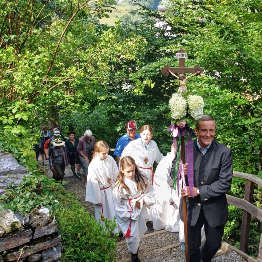 Eine Gruppe von Menschen in weißen Gewändern geht in einem Wald die Stufen hinauf. Ein Mann hält ein Kreuz mit Blumen.