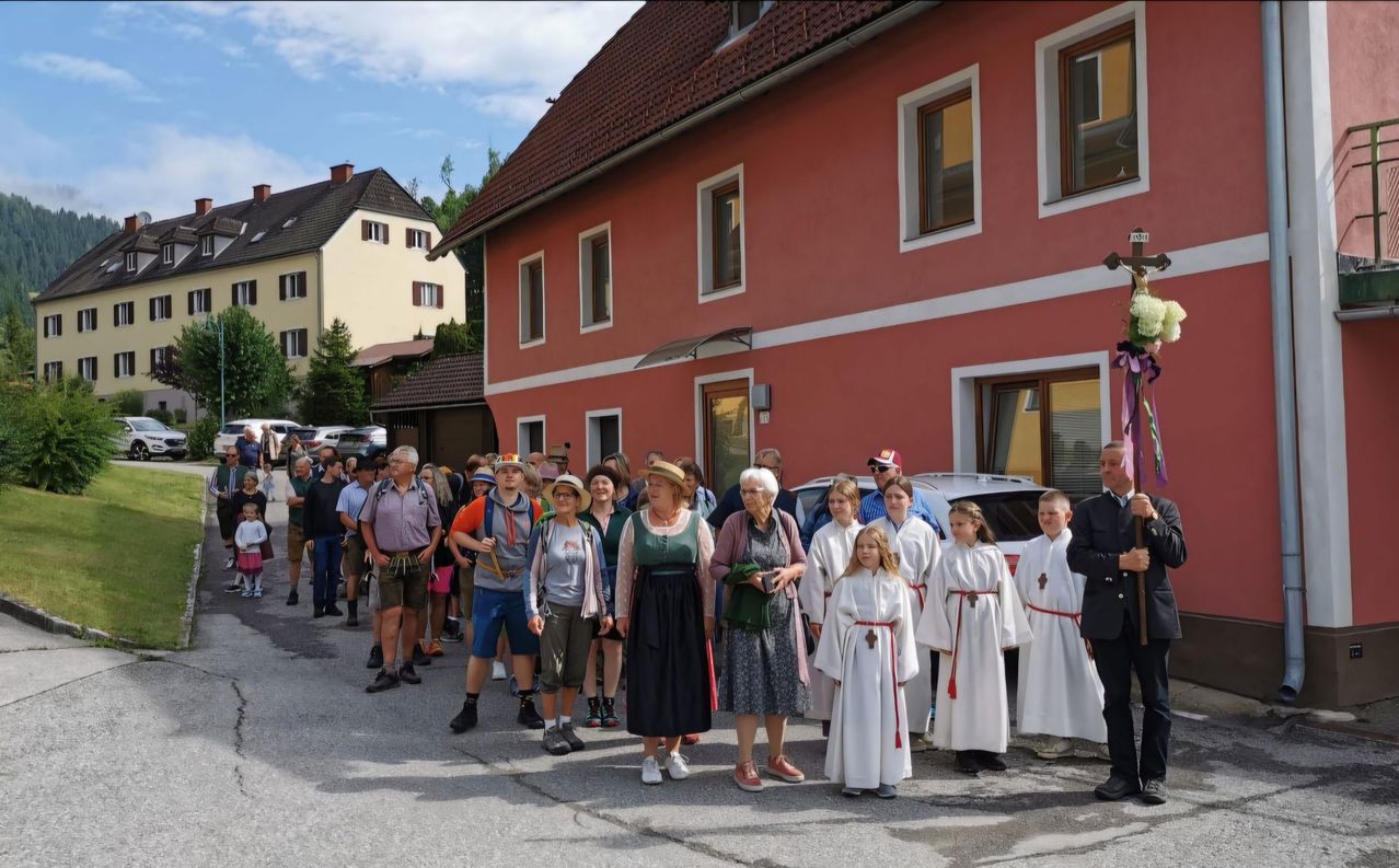 Eine Gruppe von Menschen steht in einer Reihe vor einem roten Gebäude. Einige tragen traditionelle Kleidung, andere haben Hüte. Ein Mann in einem schwarzen Anzug steht vor ihnen.