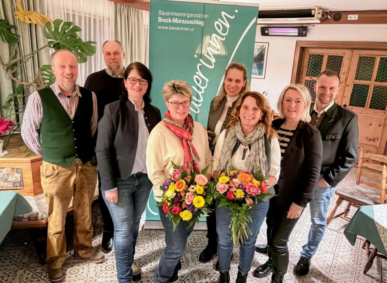 A group of adults stands in front of a banner. Two women hold flower bouquets, and the others smile. They appear to be celebrating an event.