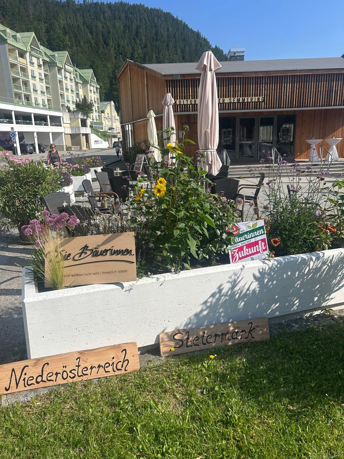 Ein Garten mit verschiedenen Blumen und Pflanzen, einschließlich Sonnenblumen, steht vor einem Gebäude mit einem Schild, das 'Steiermark' besagt. Es gibt auch Schilder, die 'Niederosterreich' und 'Die Bauern' besagen. Es gibt Tische und Stühle vor dem Gebäude.