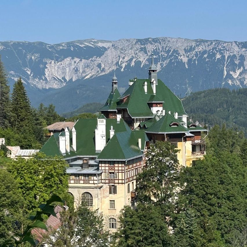 Ein großes Gebäude mit grünen Dächern und Schornsteinen, umgeben von Bäumen und Bergen im Hintergrund.