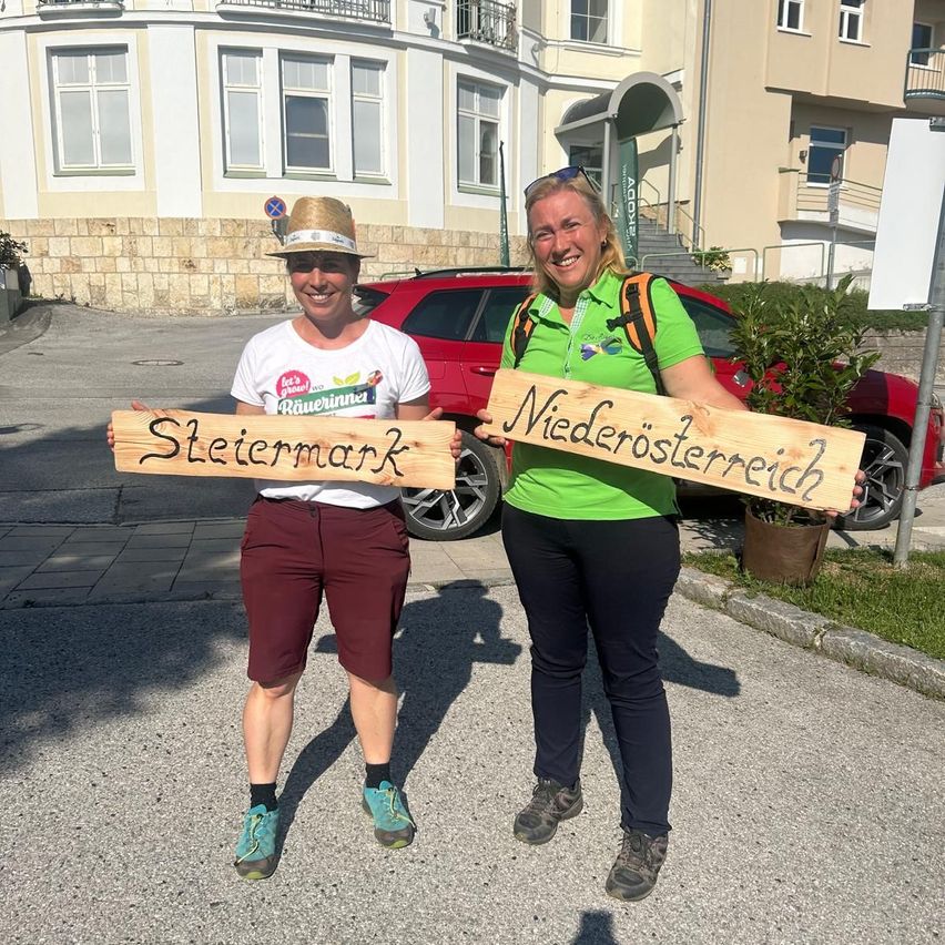 Zwei Frauen stehen auf einer Straße und halten Holzschilder mit den Aufschriften 'Steiermark' und 'Niederosterreich' in der Hand, lächeln und posieren für ein Foto. Hinter ihnen ist ein rotes Auto und ein Gebäude mit einer Treppe zu sehen.