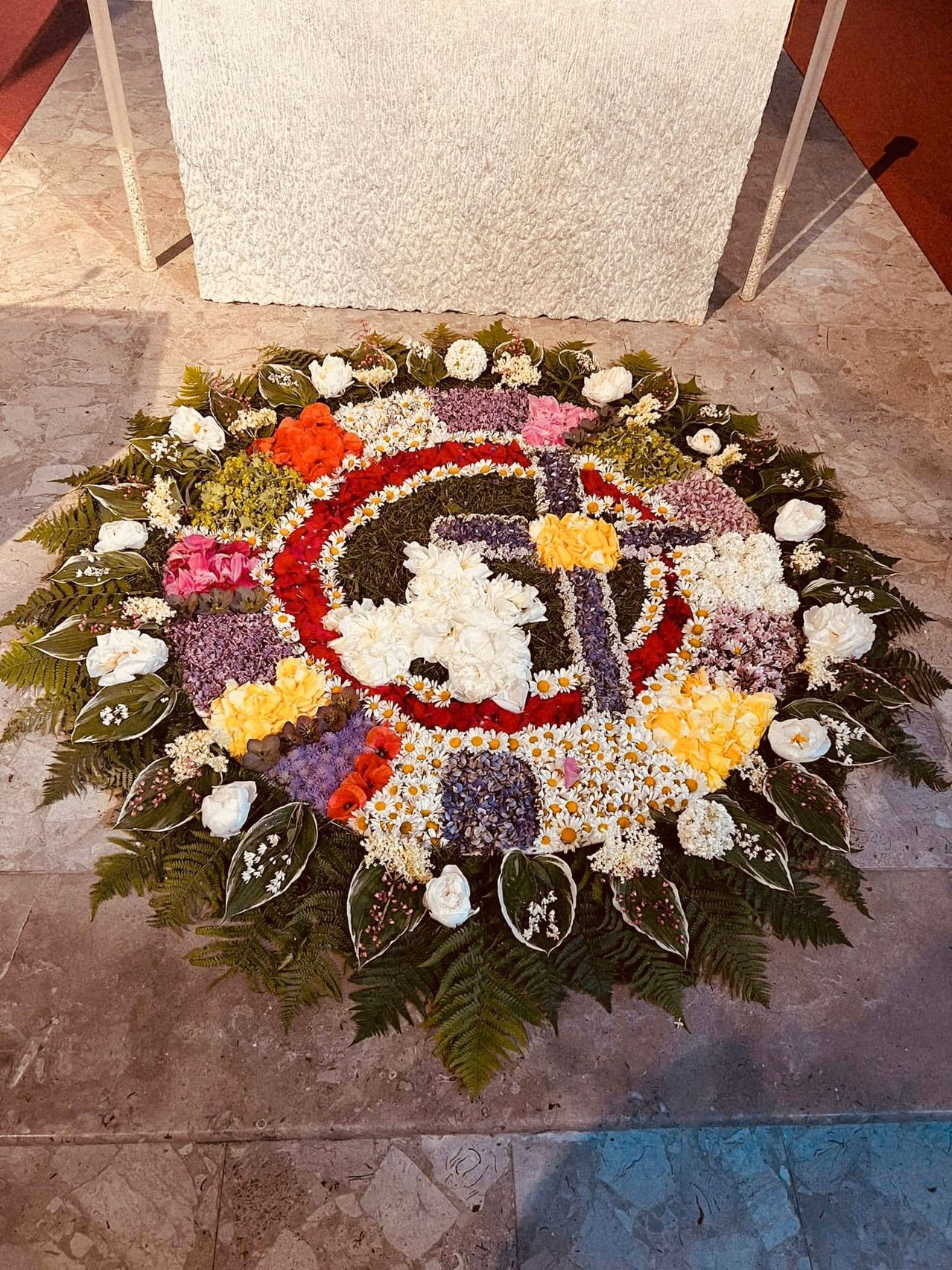A circular floral arrangement features a cross in the center, surrounded by a vibrant mix of red, white, yellow, and purple flowers, with green leaves forming the outer border.