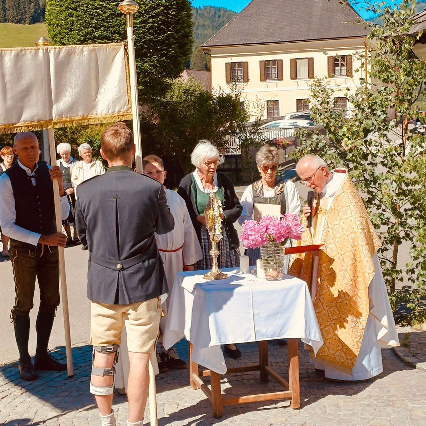 A priest stands at a table with a vase of pink flowers, surrounded by people in traditional attire, possibly at a church event.