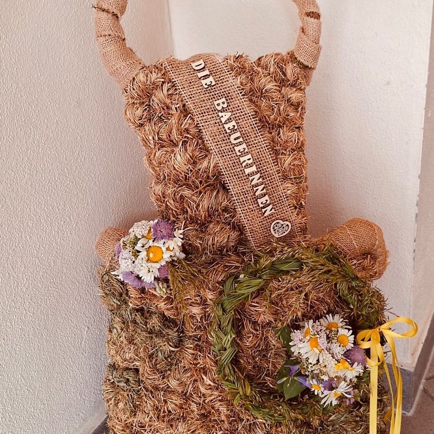 A straw bear with flowers, a sash reading 'DIE BAUERINNEN', and a yellow ribbon. The bear is standing against a light beige wall.