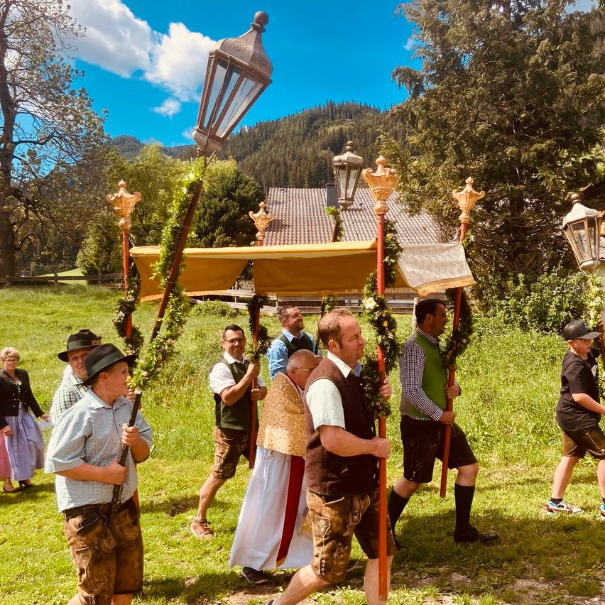 People dressed in traditional attire hold decorated poles, walking in a grassy field under a clear blue sky. A priest stands at the center, and a house with a tiled roof is behind them.