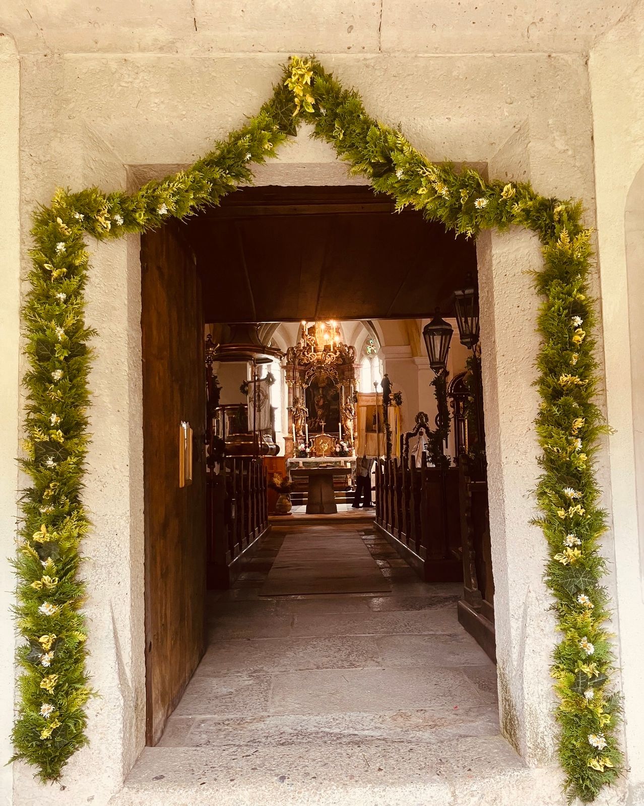 A wooden church entrance adorned with green garlands, leading to an aisle with pews. A chandelier and altar are visible at the far end.