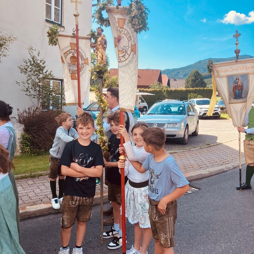 Four children in traditional German attire stand in a street, holding banners with religious symbols and a golden statue in the background. Behind them are parked cars, buildings, and mountains.