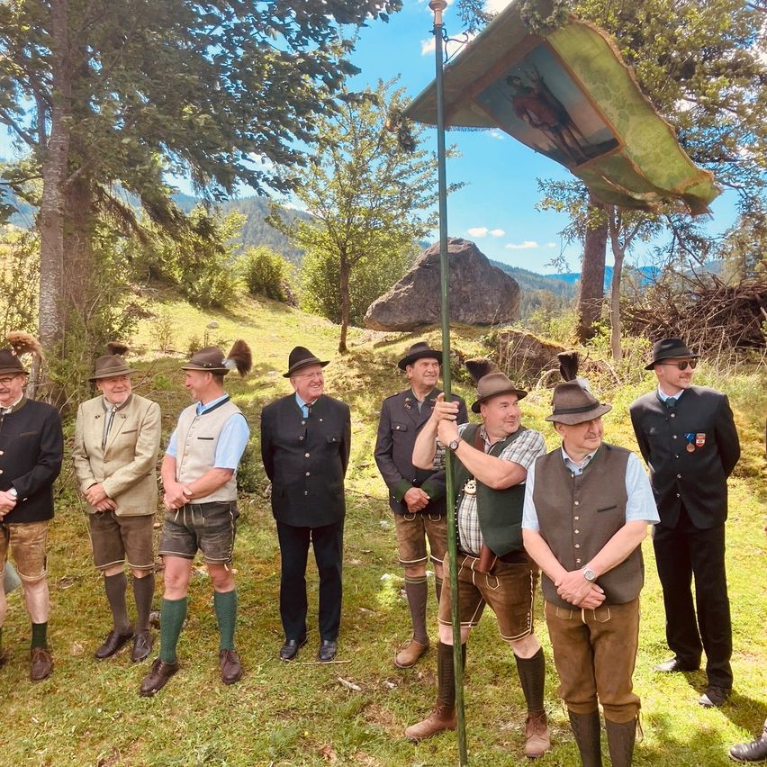 A group of men dressed in traditional clothing stands in a grassy area, one holding a flag with a historical figure. Trees and mountains in the background.