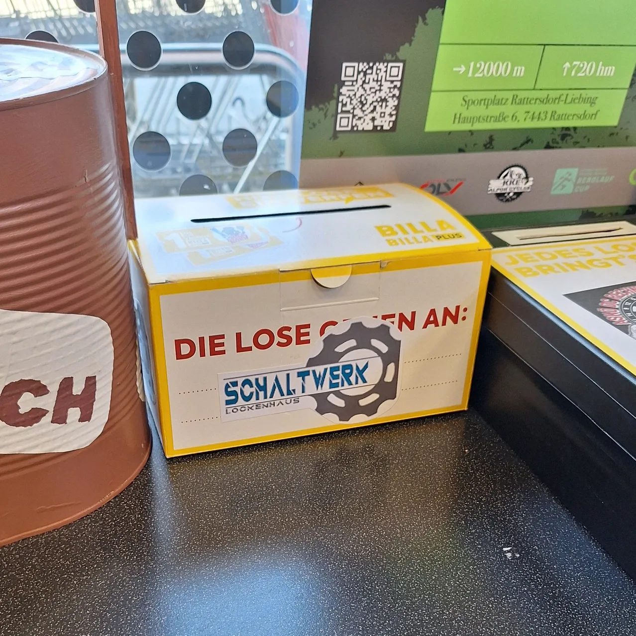 Eine gelbe Spendenbox mit dem Text "Die Lose bringen an:" steht auf einer schwarzen Oberfläche neben einer braunen Blechdose.