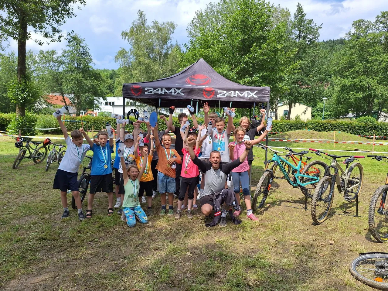 Eine Gruppe von Kindern und Erwachsenen feiert unter einem 24MX-Zelt und hält Medaillen, Wasserflaschen und Fahrräder in der Hand. Die Umgebung ist im Freien mit Bäumen und einem Gebäude im Hintergrund.