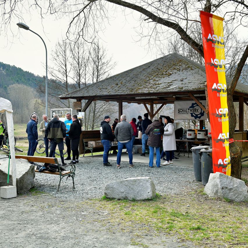 Eine Gruppe von Menschen versammelt sich unter einem Pavillon mit einem Tisch und Mülleimern in der Nähe. Ein Banner und Fahrräder sind vorhanden, und das Gebiet ist von Felsen und Bäumen umgeben.