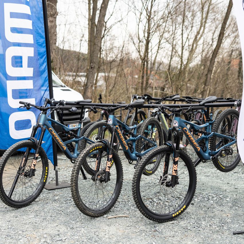 Mehrere blaue Mountainbikes sind in einem Schotterbereich geparkt. Ein blauer Banner mit weißem Text steht links. Ein weißer Van ist hinter den Fahrrädern geparkt. Im Hintergrund sind laublose Bäume zu sehen.