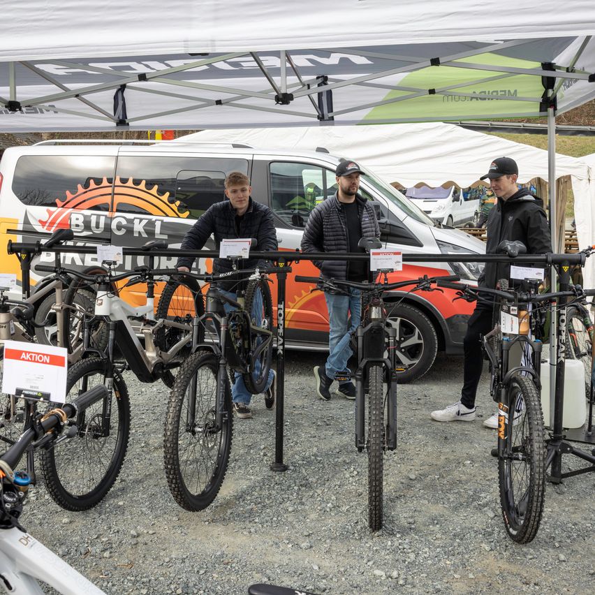 Drei Männer stehen neben einem Zelt mit mehreren ausgestellten Fahrrädern. Die Fahrräder stehen unter einem Baldachin mit dem Wort "Buckl" darauf. Sie sind auf Kiesboden geparkt.