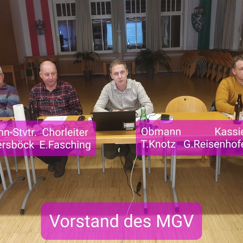 Vier Männer sitzen an einem Tisch mit Laptops, Papieren und Flaschen. Einer hat einen Namensschild mit 'Obmann'. Der Tisch hat lila Text, der 'Vorstand des MGV' sagt. Dahinter stehen Pflanzen und Stühle.