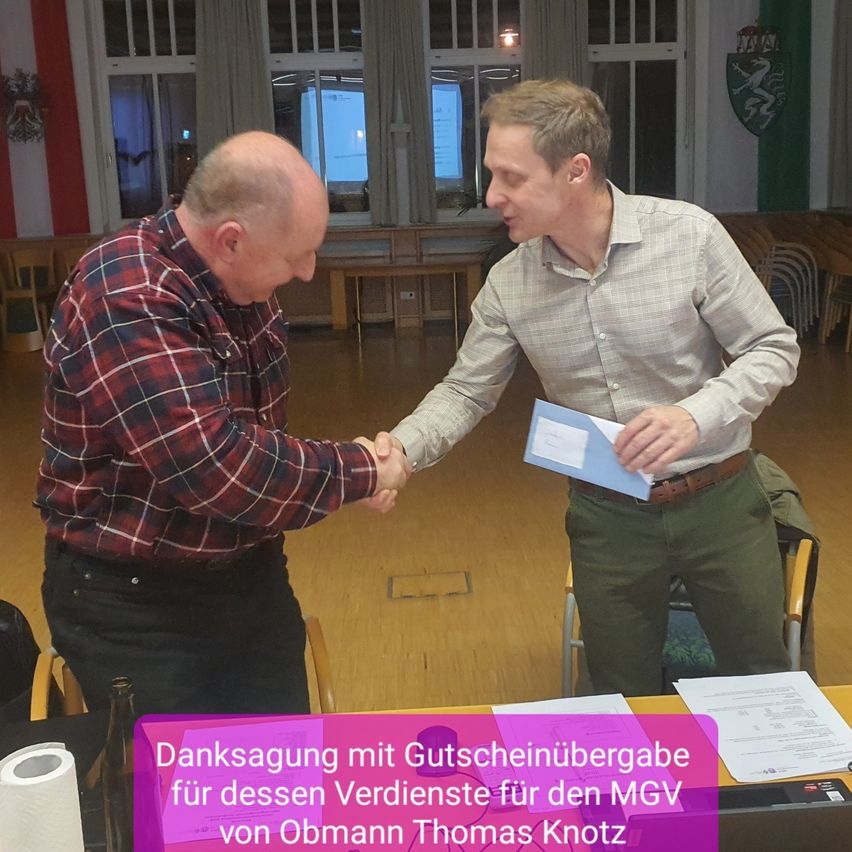 Zwei Männer schütteln sich die Hände, einer hält eine blaue Mappe, sie stehen vor einem Schreibtisch. Der Text lautet 'Danksagung mit Auszeichnungsübergabe für dessen Verdienste für den MGV von Obmann Thomas Knotz.'