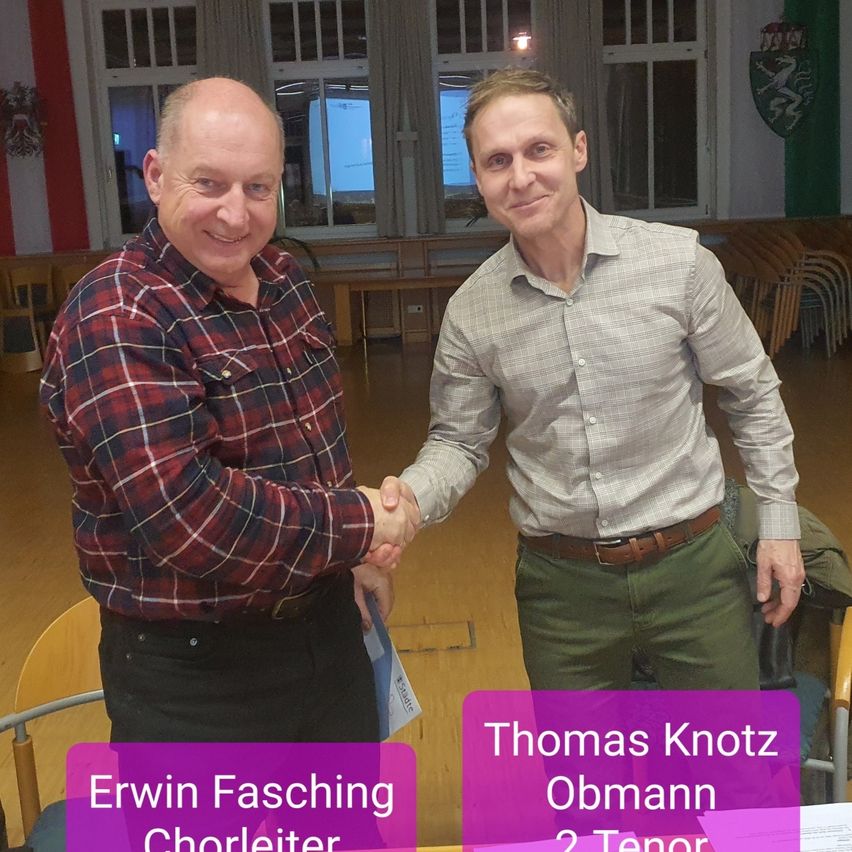 Zwei Männer stehen in einem Raum und schütteln Hände. Einer trägt ein kariertes Hemd und der andere ein hellgraues Hemd. Der Raum hat Holzböden und Stühle. Hinter ihnen befindet sich ein großes Fenster mit Vorhängen. Schilder lauten 'Erwin Fasching Chorleiter' und 'Thomas Knotz Obmann 2 Tenor.'