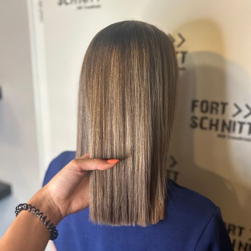 Eine Frau mit langen, gestuften braunen Haaren wird von hinten gezeigt. Sie hält ihr Haar und trägt ein blaues Oberteil. Hinter ihr befindet sich eine weiße Wand mit dem Text 'FORT SCHNITZ'.