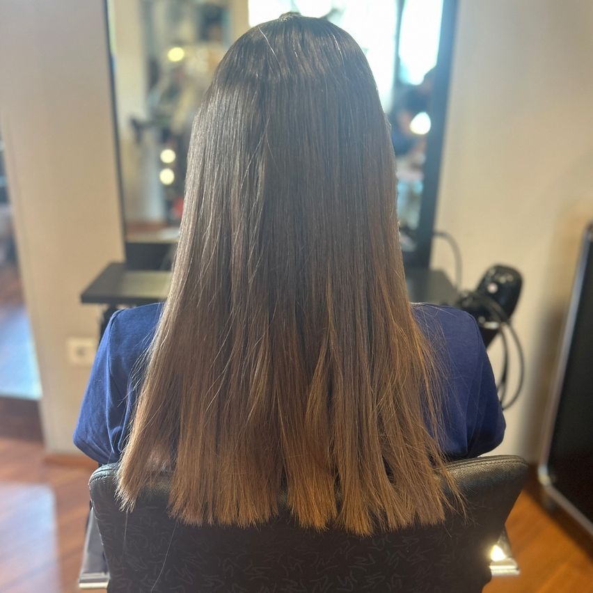 Eine Person mit langen, glatten Haaren sitzt in einem Stuhl, dem Kameraaussicht zugewendet, in einem Salon mit Spiegeln und einem Holzboden.