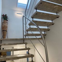 Eine moderne Treppe mit Marmorstufen und einem Metallgeländer. Eine Vase mit roten Blumen steht auf der unteren Stufe. Die Wand ist weiß und es gibt eine Deckenbeleuchtung.