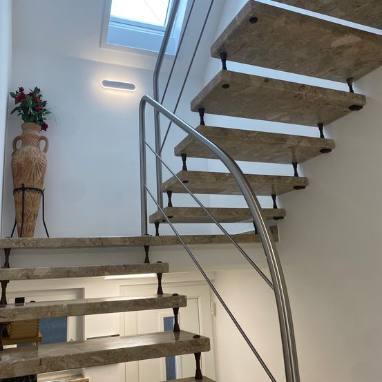 Eine moderne Treppe mit Marmorstufen und einem Metallgeländer. Eine Vase mit roten Blumen steht auf der unteren Stufe. Die Wand ist weiß und es gibt eine Deckenbeleuchtung.