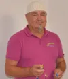 Ein Mann mit Bart und Schnurrbart trägt ein rosa Poloshirt, hält eine kleine Flasche und posiert vor einer weißen Wand.