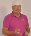 Ein Mann mit Schnurrbart und weißer Mütze lächelt und hält ein gelbes Objekt. Er trägt ein rosa Poloshirt mit Logo auf der linken Seite.