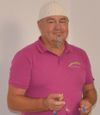 Ein Mann trägt ein rosa Poloshirt mit einem Regenbogenlogo auf der linken Brust, eine weiße Mütze und einen Schnurrbart und hält eine kleine Flasche.