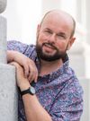 Ein Mann mit Bart und Blumenhemd lächelt, während er sich gegen eine Betonmauer lehnt.