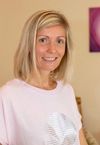 Eine Frau mit blonden Haaren lächelt und steht vor einer Wand. Sie trägt ein rosa T-Shirt mit einem weißen Design.