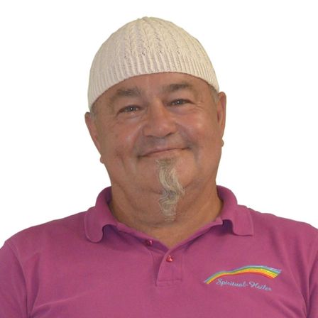 Ein lächelnder Mann mit einer weißen Strickmütze und einem Bart, der ein rosa Poloshirt mit 'Spiritueller Heiler' darauf trägt.